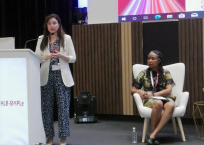 A woman stands behind a podium, speaking to a group, while another woman in a chair looks on.